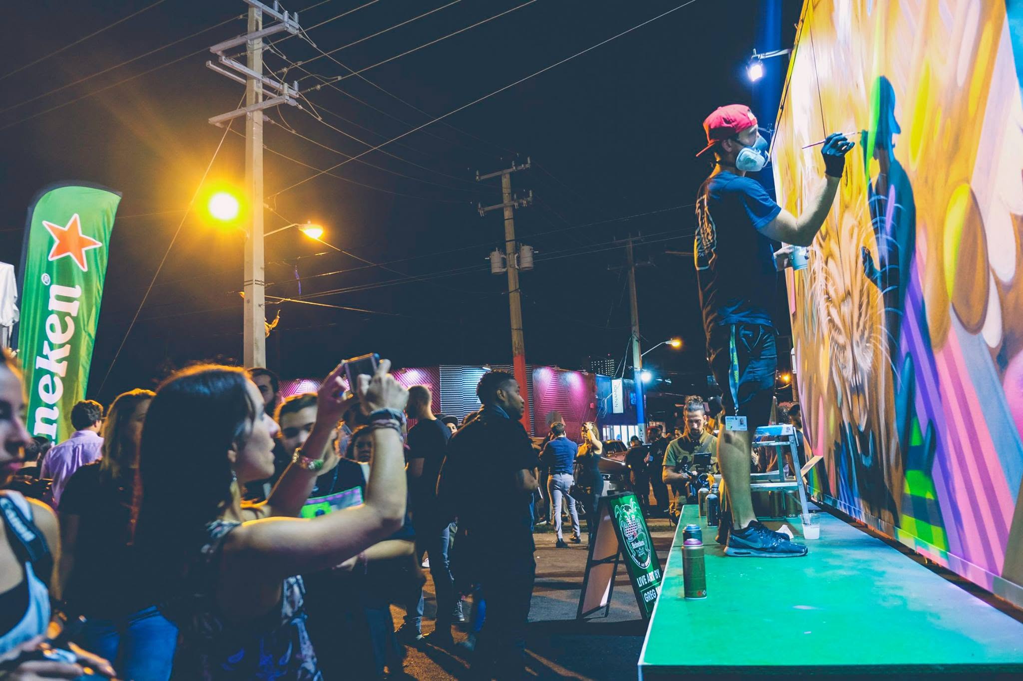 At night, a crowd beside a Heineken flag photographing an artist as he paints a bright mural on the side of a truck.