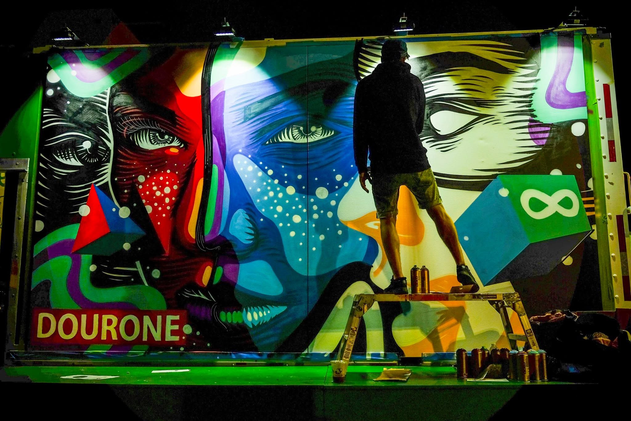 At night, an artist on a work platform spray-painting a colorful mural of stylized faces on the side of a truck.