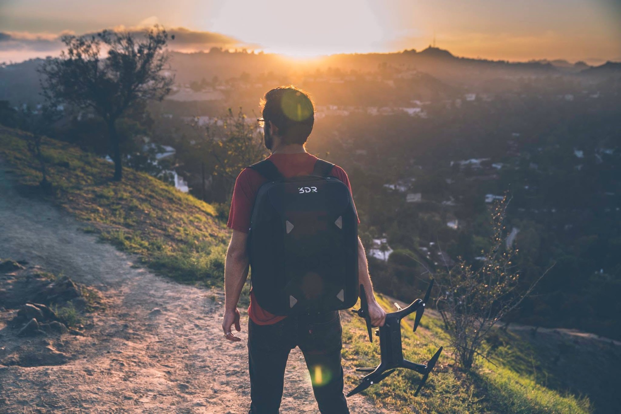 Seen from behind at sunset, a person wearing a 3DR backpack holds a drone and looks out from a hillside trail over a hazy city and hills.