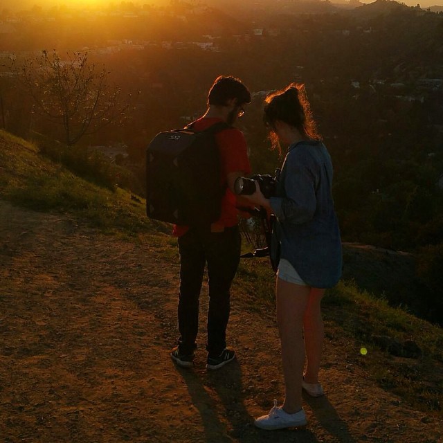At sunset on a hillside trail above the city, Kate checks a shot on her camera beside a person wearing a 3DR backpack.