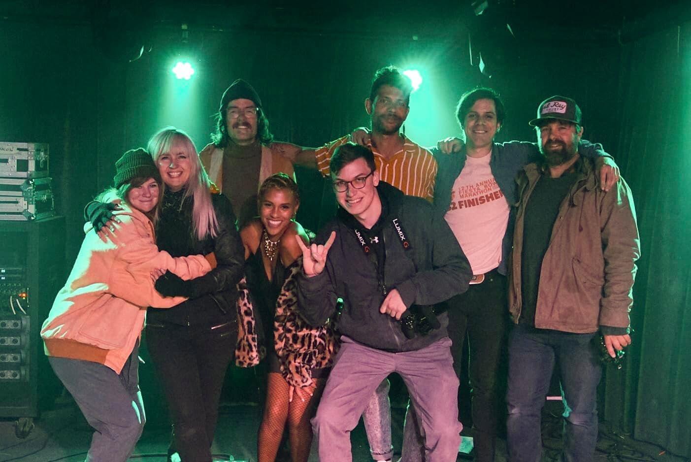 Kate, at far left, hugging Leslie Sisson of the Austin band Moving Panoramas on a dark stage lit with green lights, with others from the group smiling beside them.